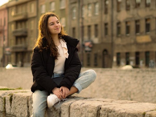 Relaxed young woman with long hair sitting on a stone wall in a cityscape.