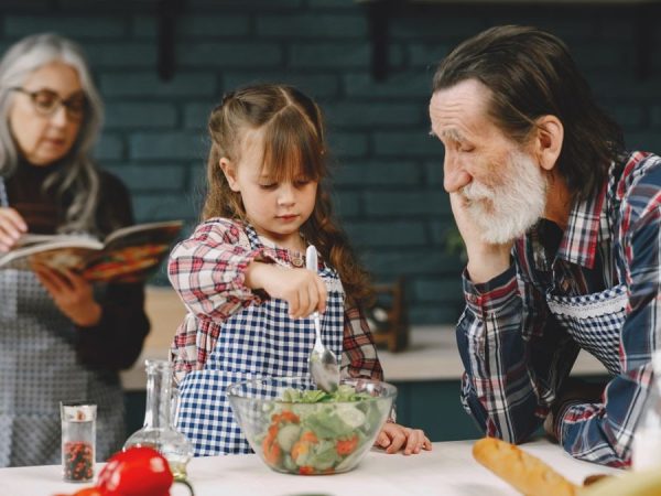 Grandfather and granddaughter making salad while grandmother reads a recipe.